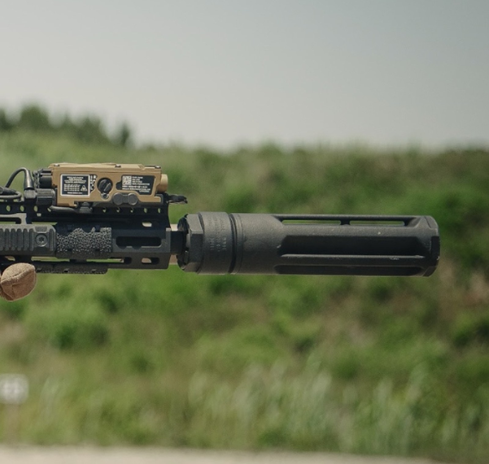 Muffler Brothers Cloak suppressor mounted to a rifle, full-body profile with mount, faceted titanium body, and muzzle brake visible against an open field